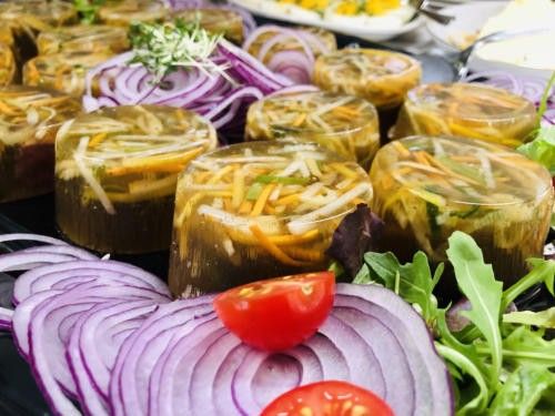 Ein schwarzer Teller mit klaren Glasbechern, gefüllt mit eingelegtem Gemüse, garniert mit Rucola, roten Zwiebeln und Tomaten. Das Gemüse ist fein gehackt und in einer klaren Flüssigkeit eingetaucht.
