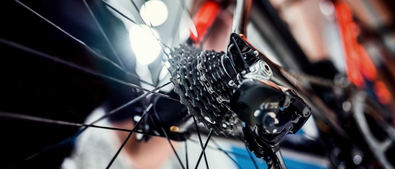 Nahaufnahme eines Hinterrad-Ritzels eines Fahrrads, das von einer Person mit Handschuhen eingestellt wird. Das Ritzel hat mehrere Zähne und ist mit der Kette verbunden. Die Hände der Person sind verschwommen.