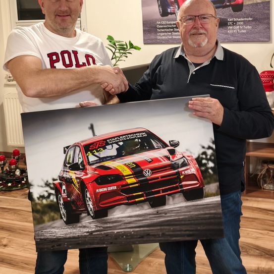 Zwei Männer halten ein gerahmtes Foto eines roten Rennwagens mit gelben und schwarzen Streifen, gekennzeichnet mit 'S13'. Sie stehen in einem Raum mit Holzboden und weißer Wand.