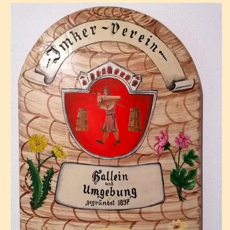 Eine Holztafel mit einem Schild zeigt eine historische Figur mit einem Stock und einem Hut, umgeben von Blumen. Die Tafel ist mit 'Ballein und Umgebung' beschriftet und wurde 1897 gegründet.