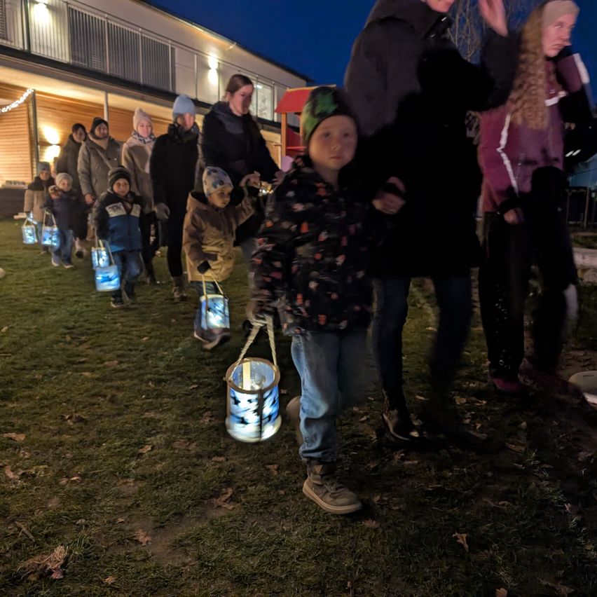 Eine Gruppe von Kindern mit leuchtenden Laternen läuft nachts einen Rasenweg hinunter, begleitet von Erwachsenen. Die Erwachsenen sind warm gekleidet und einige tragen Mützen. Die Gebäude im Hintergrund sind beleuchtet.