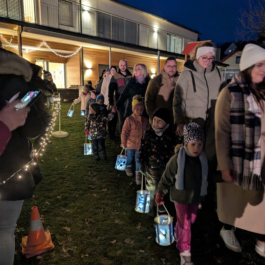 Bei Nacht laufen eine Gruppe von Menschen und Kindern mit Laternen auf einem Rasenfeld, möglicherweise für ein Festival. Ein Gebäude mit Lichtern steht hinter ihnen.