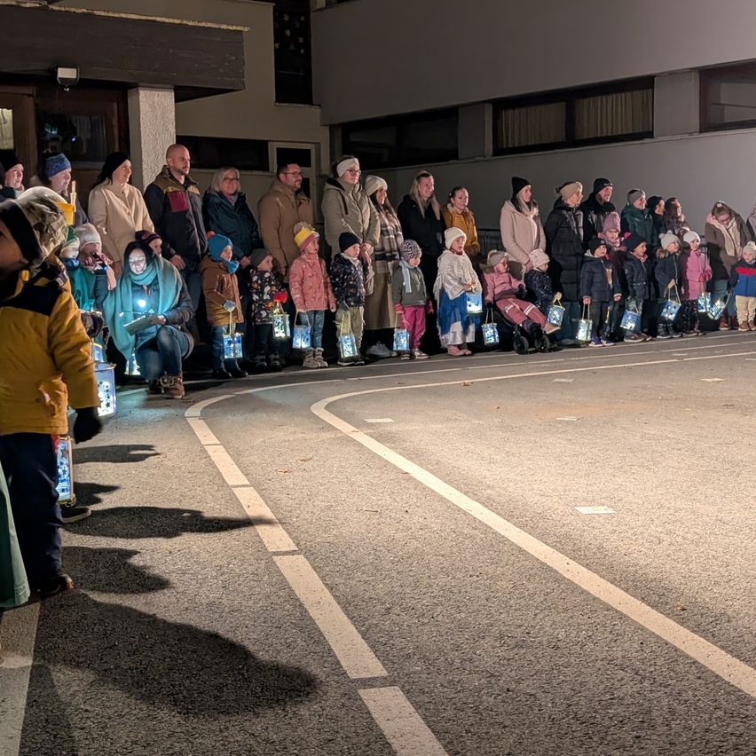 Bei Nacht steht eine Menschenmenge, einschließlich Kinder, auf der Straße und hält Laternen. Sie scheinen an einer festlichen Veranstaltung teilzunehmen.