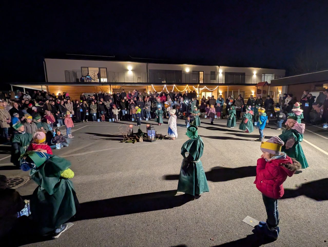 Bei Nacht versammelt sich eine große Gruppe von Menschen in einem Außenbereich, vielleicht zu einer Feier. Einige stehen und andere sitzen. Ein paar Leute tragen grüne Umhänge. Es gibt Gebäude mit Lichtern an den Seiten.
