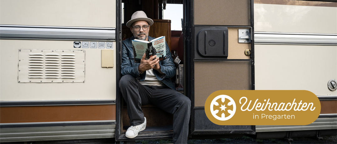 Ein älterer Mann sitzt in einem Wohnmobil und liest ein Buch mit dem Titel 'Ende gut, alles gut'. Er trägt eine Mütze und eine Brille.