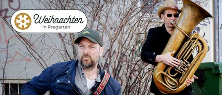 Ein Mann mit Bart und grüner Mütze steht mit einer Gitarre in einer städtischen Umgebung. Ein Schild mit der Aufschrift 'chten' ist sichtbar. Ein weiterer Mann steht neben ihm.