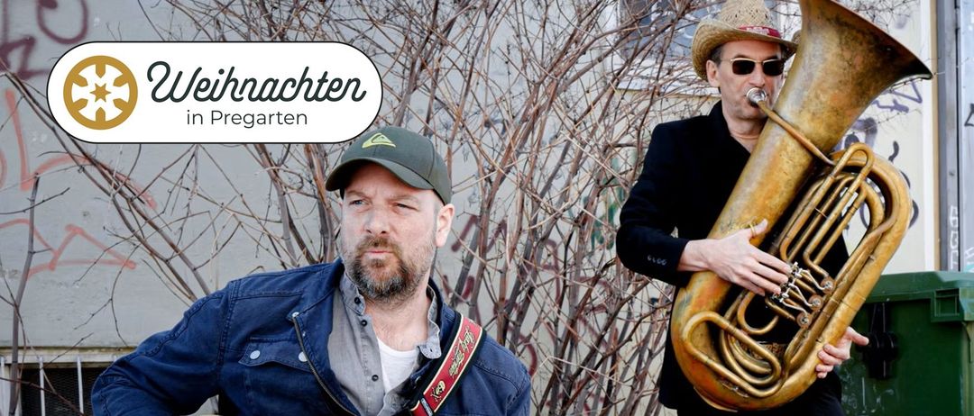 Ein Mann mit Bart und grüner Mütze steht mit einer Gitarre in einer städtischen Umgebung. Ein Schild mit der Aufschrift 'chten' ist sichtbar. Ein weiterer Mann steht neben ihm.