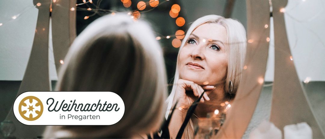 Eine Frau mit blonden Haaren schaut in einen Spiegel mit der Hand am Kinn. Dahinter sind verschwommene Lichter.