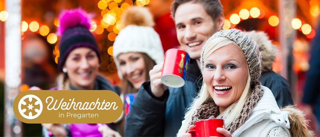 Eine Gruppe von Menschen, darunter zwei Paare, lächeln und halten rote Tassen. Sie befinden sich wahrscheinlich auf einem Weihnachtsmarkt. Der Hintergrund ist verschwommen mit Lichtern.