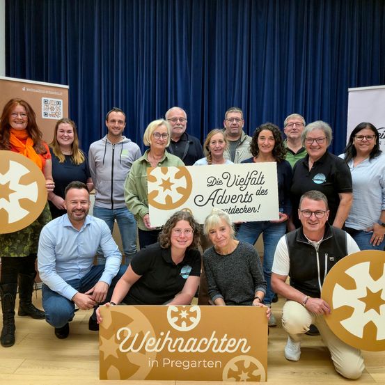 Eine Gruppe von Menschen in verschiedenen Kleidungsstücken posiert für ein Foto mit einem blauen Hintergrund. Sie halten Schilder mit den Worten 'Die Vielfalt des Advents entdecken!' und 'Weihnachten in Pregarten'.