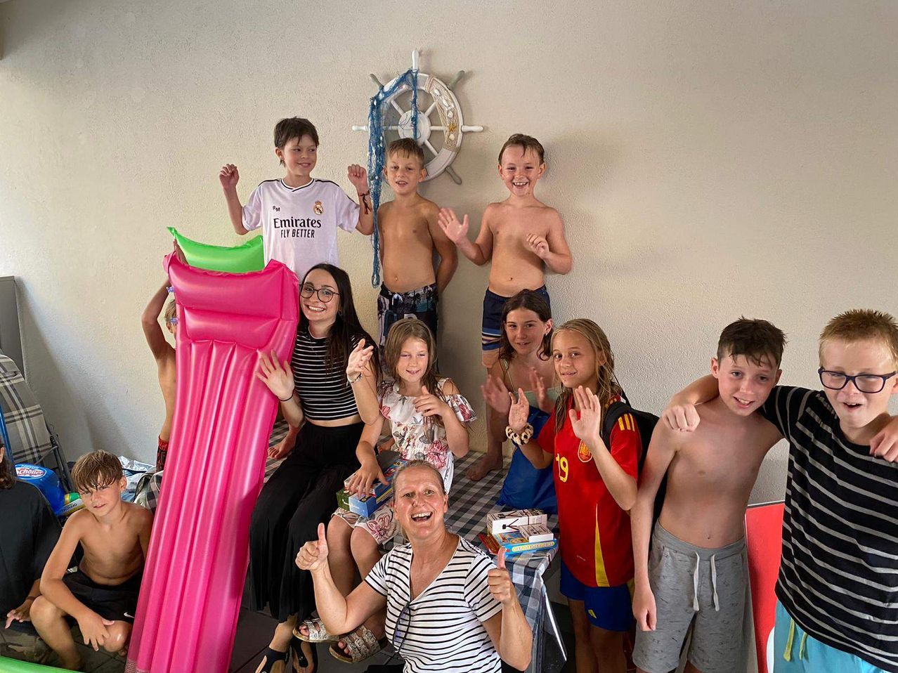 Eine Gruppe von Kindern und Erwachsenen posiert für ein Foto in einem Raum, einige stehen auf einem aufblasbaren Bett, eine Frau mit Brille sitzt, und ein dekoratives Rad an der Wand.