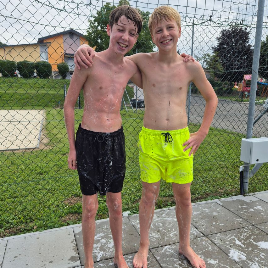 Zwei Jungen stehen nebeneinander draußen, beide mit Wasser auf dem Körper. Sie lächeln und posieren für ein Foto. Hinter ihnen steht ein Maschendrahtzaun, mit einem Rasenbereich und einem Haus in der Ferne.