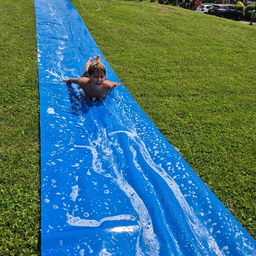 Ein junger Junge rutscht auf einem blauen Plastikrutscher auf einem Rasenfeld und hinterlässt Wasser.