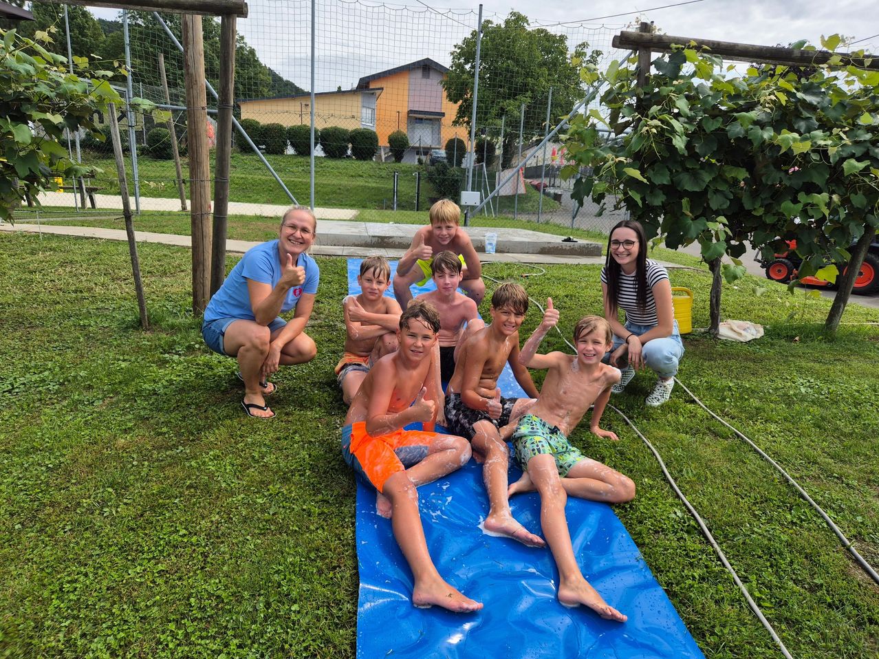 Eine Gruppe junger Jungen und Mädchen mit zwei Frauen, eine mit einem gelben Eimer, spielt auf einem blauen Wasserrutsche auf einem Rasen und zeigt Daumen hoch.