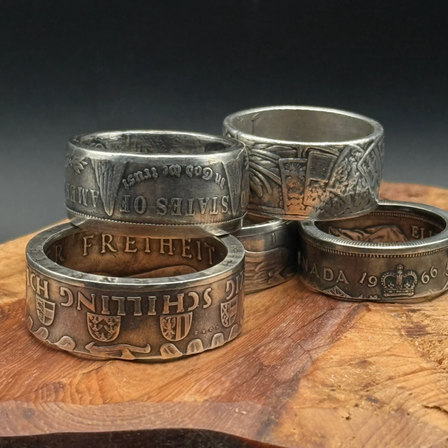 Vier silberne Ringe mit aufwendigen Verzierungen liegen auf einer Holzoberfläche. Jeder Ring hat einzigartige Gravuren wie 'FREIHEITS', 'E', 'SOVEREIGNTY' und 'CHURCH OF CHRIST'. Ein Ring hat eine Krone und 'CANADA 1966'.