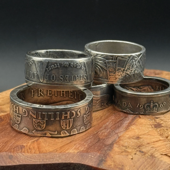 Vier silberne Ringe mit aufwendigen Verzierungen liegen auf einer Holzoberfläche. Jeder Ring hat einzigartige Gravuren wie 'FREIHEITS', 'E', 'SOVEREIGNTY' und 'CHURCH OF CHRIST'. Ein Ring hat eine Krone und 'CANADA 1966'.