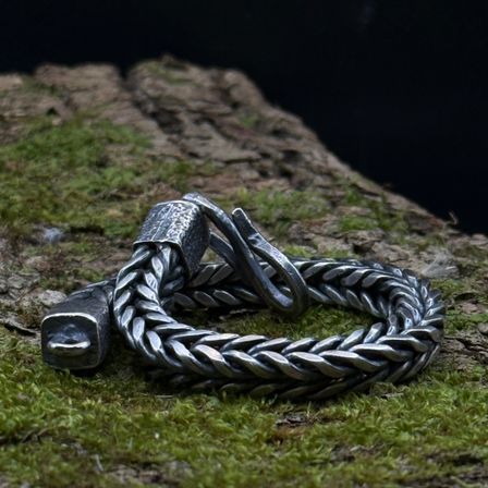 Ein silbernes Armband mit zwei Verschlüssen liegt auf einem moosbedeckten Baumstumpf. Die Kette hat eine gewebte Textur, und die Verschlüsse sind leicht gebogen.