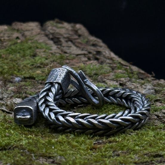 Ein silbernes Armband mit zwei Verschlüssen liegt auf einem moosbedeckten Baumstumpf. Die Kette hat eine gewebte Textur, und die Verschlüsse sind leicht gebogen.