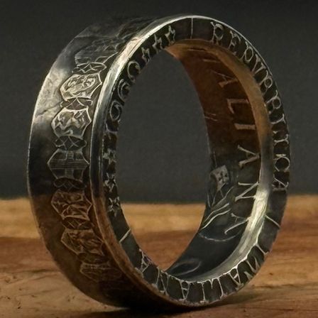 Ein silberner Ring mit kunstvollen Schnitzereien und einem braunen Zentrum, versehen mit Text und Designs, ruht auf einer Holzoberfläche.