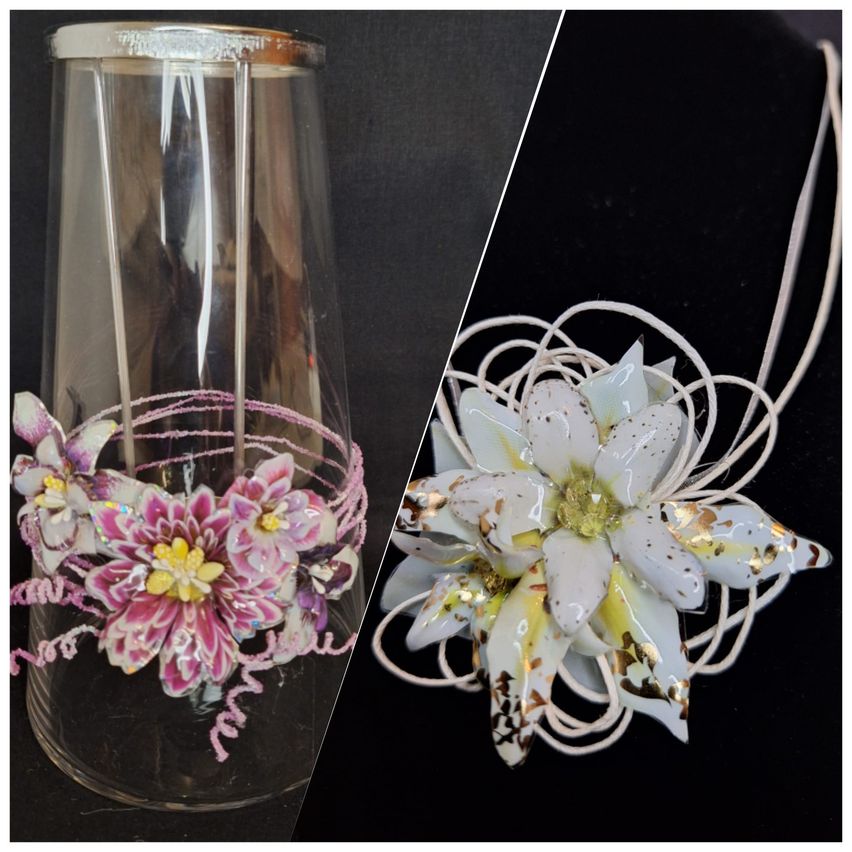 A transparent glass vase with a floral arrangement of pink and yellow flowers on the side. A white flower brooch with gold accents is placed on a black background.