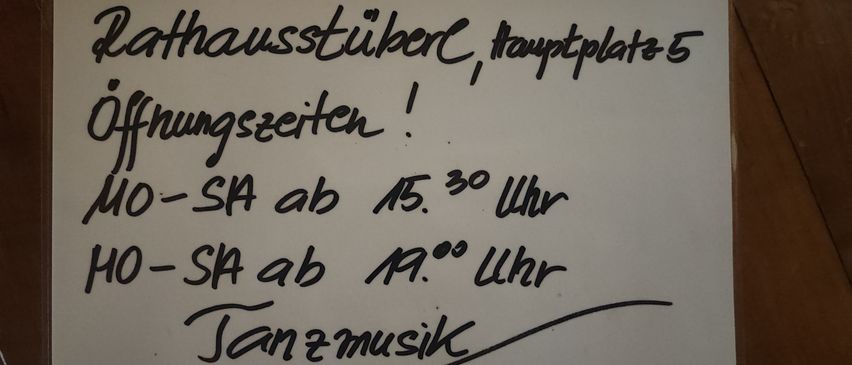 Eine handgeschriebene Notiz auf einem weißen Papier mit schwarzer Tinte. Es steht: Rathausstuhler, Hauptplatz 5, Öffnungszeiten: 10-SA ab 15.30 Uhr, 10-SA ab 19.00 Uhr, Tanzmusik.