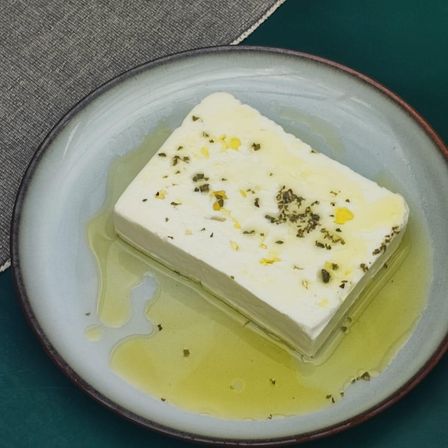 Ein würfelförmiges Stück Feta-Käse liegt auf einem Teller, umgeben von Kräutern und Olivenöl. Der Käse hat eine cremige Textur und ist mit einigen verstreuten Kräutern garniert.