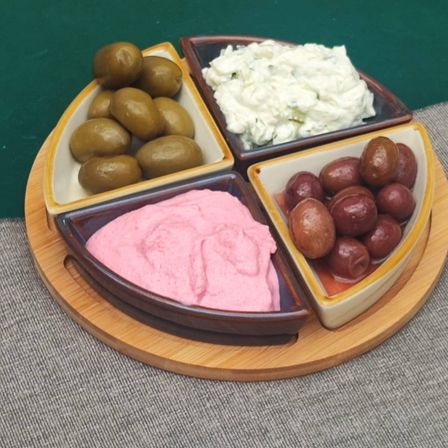 Ein Holztablett zeigt vier kleine Keramikschalen mit verschiedenen Dips und Oliven. Eine Schüssel enthält rosa Dip, eine andere grüne Oliven, die dritte Schüssel hat weißen Dip und die letzte Schüssel hat rote Oliven.