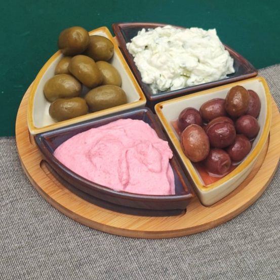Ein Holztablett zeigt vier kleine Keramikschalen mit verschiedenen Dips und Oliven. Eine Schüssel enthält rosa Dip, eine andere grüne Oliven, die dritte Schüssel hat weißen Dip und die letzte Schüssel hat rote Oliven.