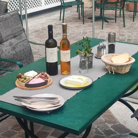 Ein grüner Außentisch mit einem Teller Käse, Oliven und einem Korb Brot, begleitet von zwei Flaschen Wein, einem Topf mit Pflanzen und Besteck.