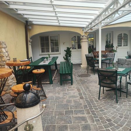 Ein Restaurant im Freien mit Tischen und Stühlen unter einem Glasdach. Die Tische haben grüne Tischdecken und einige haben Pflanzen und Tassen. Die Stühle sind grün mit Rückenlehnen. Der Boden ist aus Stein und in der Mitte stehen Säulen.