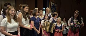 Eine Gruppe von Frauen und Mädchen in traditioneller Kleidung steht auf der Bühne und lächelt für ein Foto. Zwei Mädchen halten ein Akkordeon und alle halten kleine Geschenke. Im Hintergrund hängt ein Banner.