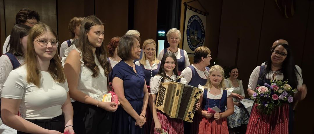 Eine Gruppe von Frauen und Mädchen in traditioneller Kleidung steht auf der Bühne und lächelt für ein Foto. Zwei Mädchen halten ein Akkordeon und alle halten kleine Geschenke. Im Hintergrund hängt ein Banner.