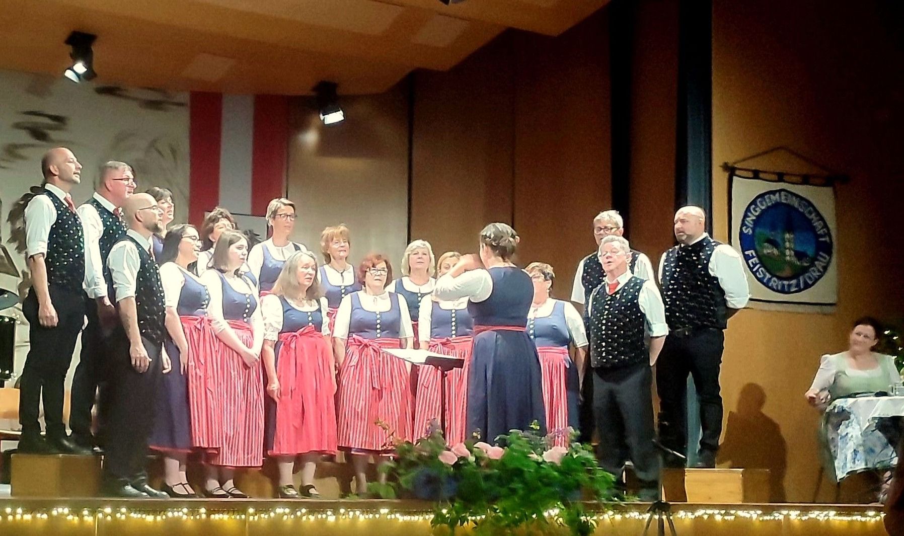 Ein Chor in traditioneller Kleidung tritt auf der Bühne auf. Einige Mitglieder tragen rot-weiß gestreifte Kleider, andere blau-weiße. Ein Mann und eine Frau stehen vorne, mit einer Lampe über ihnen.