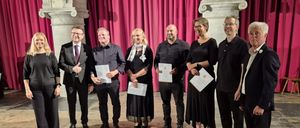 Eine Gruppe von sechs Personen steht auf einer Bühne mit rotem Vorhang im Hintergrund und hält Urkunden in den Händen. Sie tragen formelle Kleidung und lächeln, möglicherweise zur Feier einer Leistung.