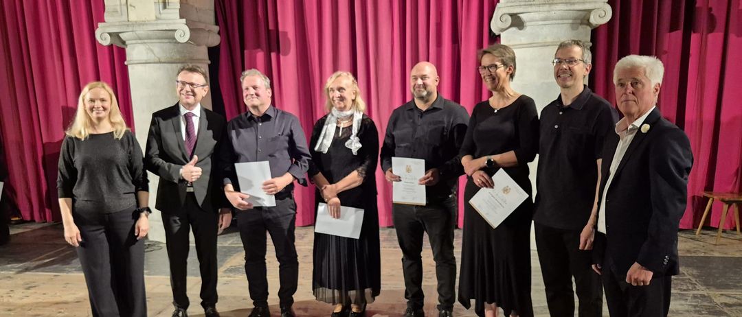 Eine Gruppe von sechs Personen steht auf einer Bühne mit rotem Vorhang im Hintergrund und hält Urkunden in den Händen. Sie tragen formelle Kleidung und lächeln, möglicherweise zur Feier einer Leistung.