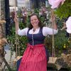 Eine Frau in traditioneller Kleidung steht auf einer dekorativen Schaukel, geschmückt mit großen rosa Blumen, und posiert für ein Foto vor einem Gebäude.