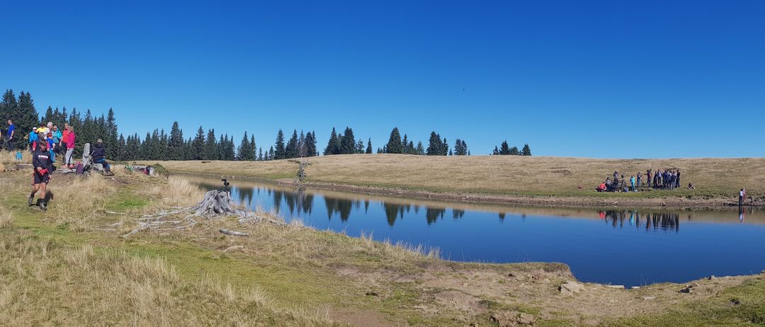 Eine Gruppe von Menschen versammelt sich auf einem Grasfeld mit einem See davor. Das Feld ist von Bäumen umgeben, und der Himmel ist klar und blau.