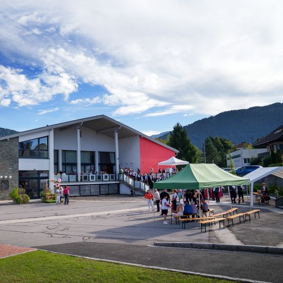 Ein großes Gebäude mit einer rot-weißen Flagge davor. Mehrere Menschen sind draußen versammelt, einige unter einem grünen Zelt. Im Hintergrund befinden sich Berge und ein blauer Himmel mit Wolken.