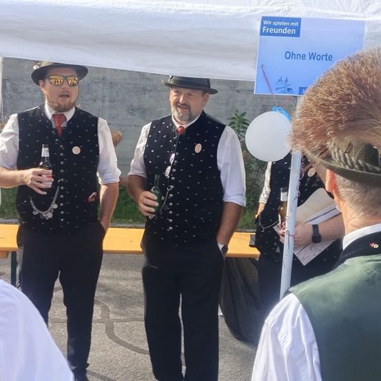 Zwei Männer in traditioneller Kleidung stehen unter einem Zelt, halten Getränke und lesen Schilder 'Wir spielen mit Freunden' und 'Ohne Worte'. Ein Mann mit Fellmütze beobachtet sie.