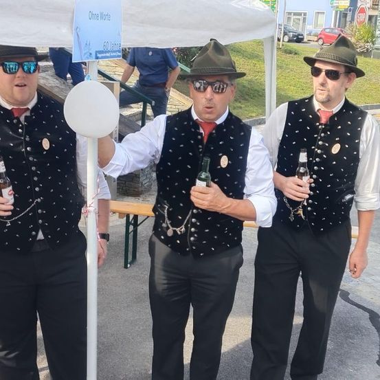 Drei Männer stehen draußen mit Bierflaschen. Zwei tragen Sonnenbrillen und Hüte. Ein Schild hinter ihnen steht Ohne Worte. Ein Stoppschild und geparkte Autos sind im Hintergrund sichtbar.