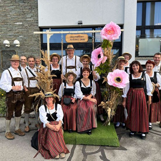 Eine Gruppe von Menschen in traditioneller Kleidung steht vor einem Gebäude. Einige sitzen auf einem grünen Teppich, während andere stehen. Sie alle lächeln und posieren für ein Foto. Das Gebäude hat ein Holzschild mit Text.