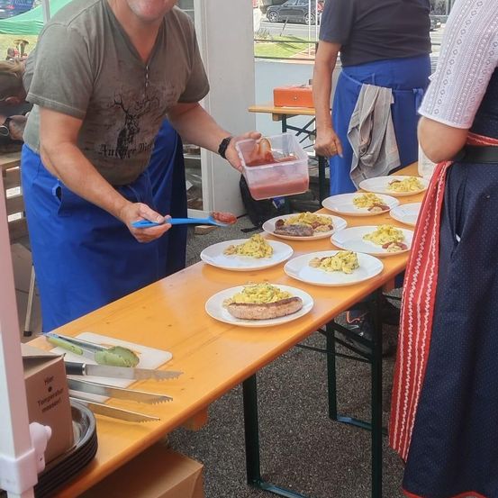 Ein Mann in einem braunen T-Shirt und blauer Hose serviert Essen auf einem Holztisch. Eine weitere Person in einer blauen Schürze steht neben ihm. Der Tisch ist mit Tellern gefüllt. Ein Küchenwerkzeug und eine Box stehen auf dem Tisch. Im Hintergrund stehen Leute.