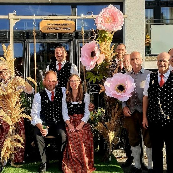 Eine Gruppe von Menschen posiert für ein Foto vor einem Gebäude mit rosa Blumen und einem Schild, das Feiertriz/Drau besagt.