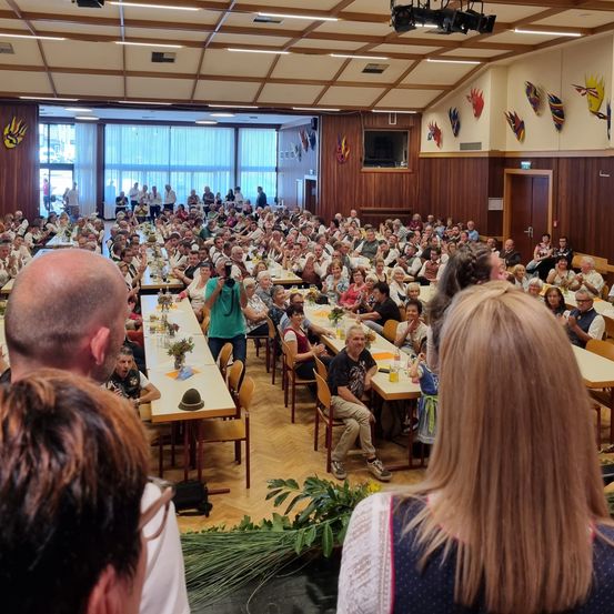 Ein großer Innenraum mit Holzwänden und hoher Decke. Viele Menschen sitzen an langen Tischen, einige klatschen. Im Vordergrund befindet sich eine Bühne mit Mikrofon und Pflanzen.