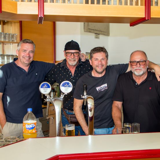 Vier Männer stehen hinter einer Bar und lächeln für ein Foto. Sie tragen schwarze T-Shirts, eines mit einem Levis-Logo. Bierhähne und Gläser stehen auf der Bar, neben einer Fanta-Flasche. Regale im Hintergrund halten Gläser.