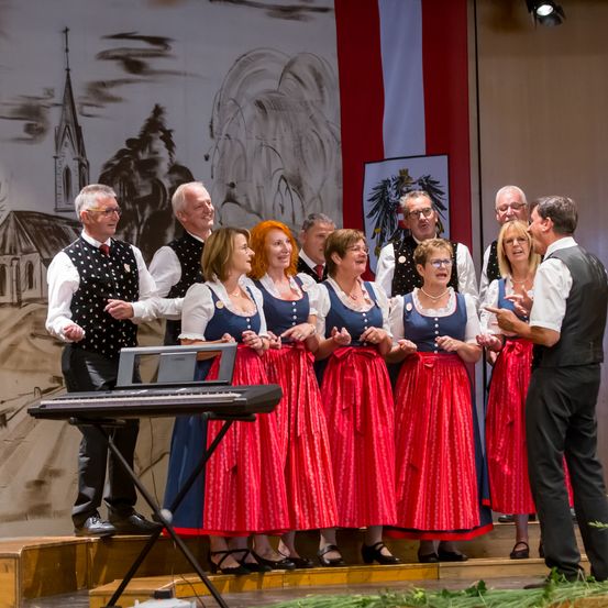 Eine Gruppe von Menschen in traditioneller Kleidung tritt auf der Bühne auf, mit einem Klavier vor ihnen. Dahinter befindet sich eine Wandmalerei und Flaggen.