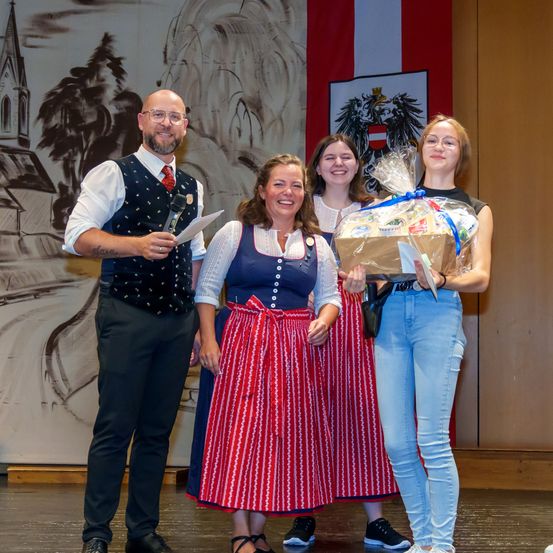 Vier Personen auf der Bühne; eine hält ein Mikrofon, eine andere einen verpackten Korb und zwei Frauen lächeln. Im Hintergrund befindet sich ein Wandbild einer Kirche und Bäume.