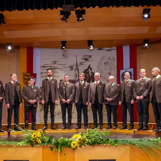 Eine Gruppe von Männern in braunen Anzügen mit roten Krawatten steht auf der Bühne. Sie haben Blumen vor sich auf der Bühne und Scheinwerfer an der Decke.