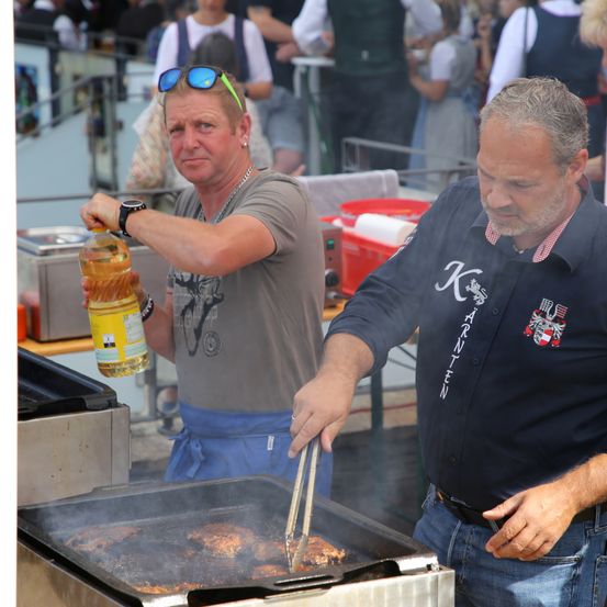 Zwei Männer braten Essen auf einem Grill. Einer gießt Öl auf den Grill, während der andere das Essen mit einer Zange wendelt. Der Mann auf der linken Seite trägt ein graues T-Shirt, während der Mann auf der rechten Seite ein blaues Hemd mit Logo trägt.