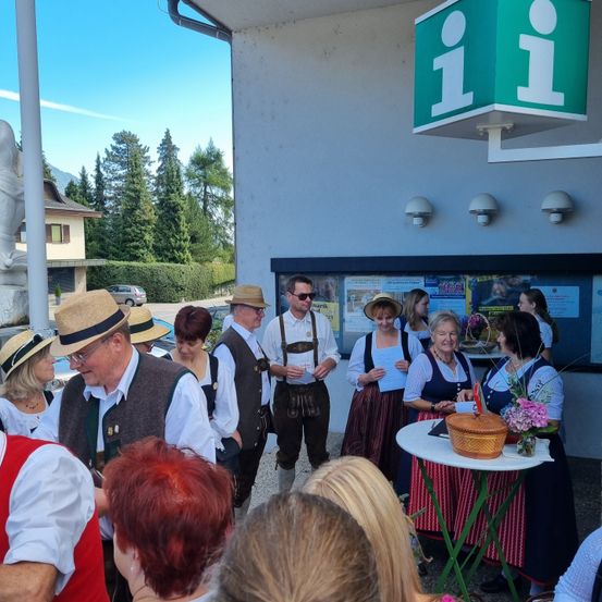 Eine Gruppe von Menschen in traditioneller Kleidung steht vor einem Gebäude, möglicherweise für eine kulturelle Veranstaltung. Ein Tisch mit Blumen und einem Korb ist aufgestellt.